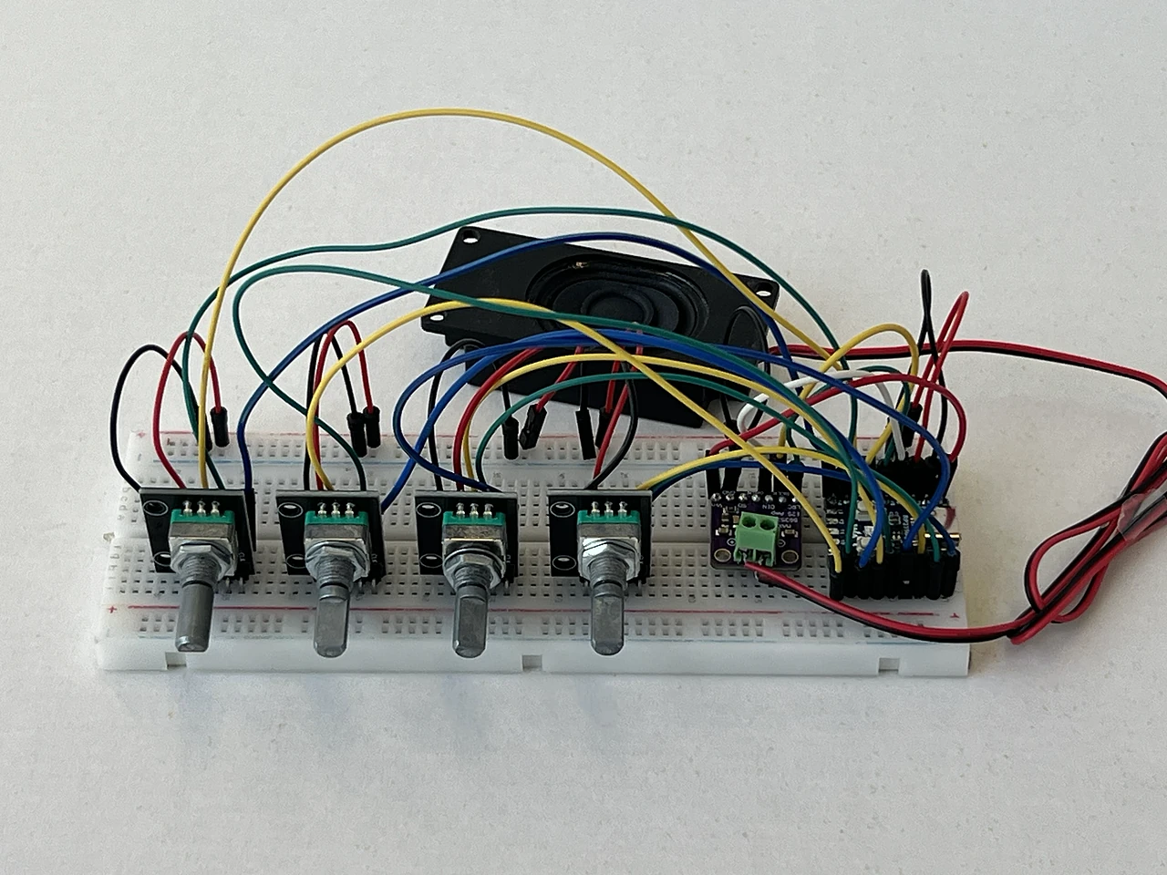 Das fertige Gerät von vorn: die vier Regler stehen mit ihren Metallachsen nebeneinander an der Vorderkante des Steckbretts, dahinter liegt der schwarze Lautsprecher, rechts daneben sitzen Audioboard und Chip. Über allem spannen sich gelbe, grüne, blaue, rote und schwarze Kabel.